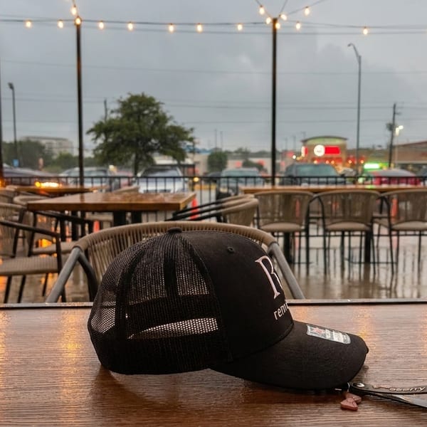 Rainy outdoor patio with a black baseball cap on a wet wooden table, string lights overhead.